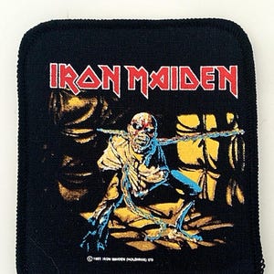 May include: A black Iron Maiden patch featuring a yellow and blue image of Eddie, the band's mascot, with the text "Iron Maiden" in red and white above him. The text "© 1987 Iron Maiden (Holdings) Ltd" is at the bottom of the patch.