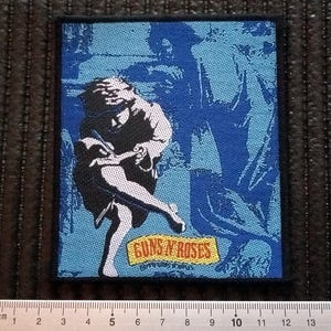 Op de afbeelding: Een blauwe en witte geborduurde patch met het Guns N' Roses logo en de tekst "Use Your Illusion I" in wit. De patch toont een silhouet van een persoon met lang haar.