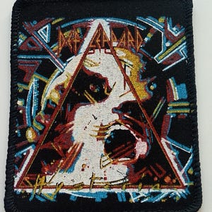 Könnte beinhalten: Schwarzer gewebter Patch mit einer roten, gelben und blauen Grafik eines schreienden Gesichts in einem Dreieck. Der Text "Megadeth" ist in einer stilisierten Schrift über dem Dreieck geschrieben.