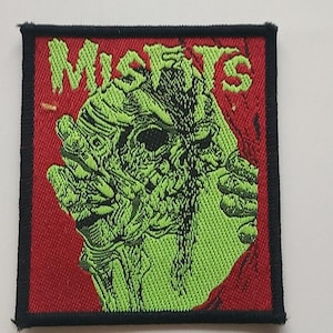 Może przedstawiać: Kwadratowa naszywka z czarną obwódką z napisem "MISFITS" w zielonych blokowych literach nad zielono-czarną grafiką twarzy przypominającej czaszkę na czerwonym tle.