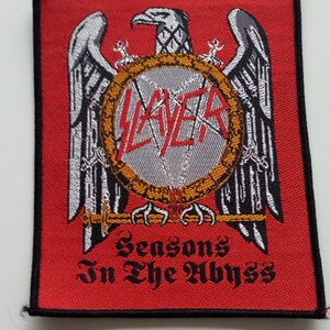 Puede incluir: Un parche bordado rojo con borde negro que presenta un águila plateada con las alas extendidas sosteniendo un emblema circular con la palabra "Slayer" en rojo y las palabras "Seasons In The Abyss" en negro.