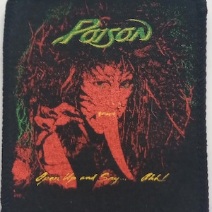 Op de afbeelding: Een zwarte stoffen patch met een groen-gele band met de tekst "FAUST" en een rood-gele afbeelding van een vrouw met een lange neus. De tekst "Open Up and Say... Ah!" is onderaan in geel gedrukt.