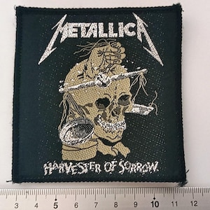 May include: Black and white embroidered patch featuring the band name "Metallica" and the text "Harvester of Sorrow". The patch depicts a skull with a silver scythe.