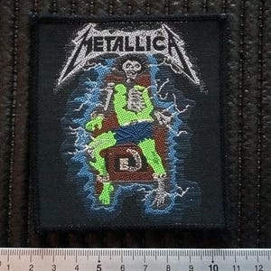 Op de afbeelding: Zwarte en witte geborduurde patch met de bandnaam "METALLICA" in witte letters. De patch toont een groen skelet dat op een troon zit met bliksemstralen erachter.
