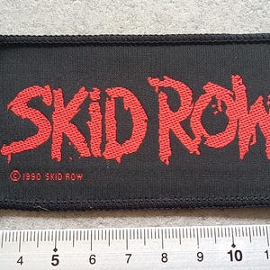 Pode incluir: Patch retangular preto com o texto vermelho "SKID ROW" em uma fonte desgastada. O texto "©1990 SKID ROW" está abaixo. O patch tem uma borda preta e está sobre uma superfície cinza. Uma régua está abaixo.