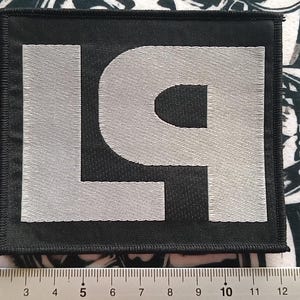 Op de afbeelding: Een vierkante zwarte patch met de geborduurde letters "L9" in zilver. De patch is geplaatst op een patroon oppervlak met een liniaal in centimeters langs de onderkant.