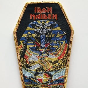 Könnte beinhalten: Ein schwarzer und goldener Bügelaufkleber mit einer Schwarzweißillustration eines Pharaos mit dem Text "Iron Maiden" in roten und goldenen Buchstaben.
