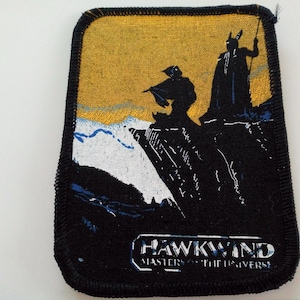 Op de afbeelding: Een zwart en geel geborduurd patch met een silhouet van twee figuren die op een klif staan. De tekst "Hawkwind Masters of the Universe" is onder de figuren gedrukt.