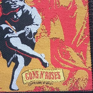 Op de afbeelding: Een rechthoekige patch met een gestileerde illustratie van een figuur met een pen, tegen een rode en gele achtergrond. De patch bevat de tekst "GUNS N' ROSES" in een banner. De patch is genaaid op een zwart oppervlak.