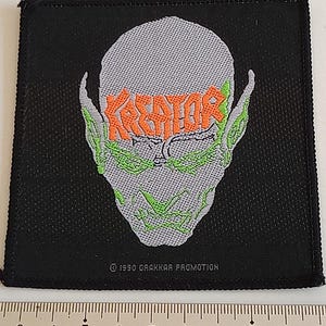 May include: Black fabric patch featuring a grey alien head with green eyes and the word "Kreator" in orange lettering. The text "© 1990 DRAKKAR PROMOTION" is at the bottom of the patch.