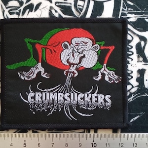 Op de afbeelding: Zwarte en groene geborduurde patch met een cartoonmonster dat groene rook blaast. De tekst "CRUMBSUCKERS" staat onder het monster.