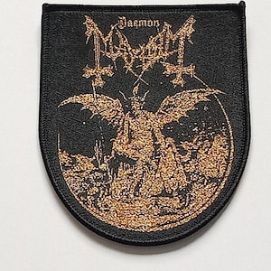 Op de afbeelding: Zwarte patch met een gouden illustratie van een gevleugelde figuur, omgekeerde kruisen en de bandnaam "Mayhem". Het woord "Daemon" staat boven de bandnaam. De patch heeft een zwarte rand en een schildvorm.