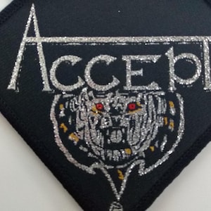 Op de afbeelding: Een zwarte stoffen patch met zilveren glitterletters die "ACCEPT" lezen. Onder de letters staat een zilveren glitterafbeelding van een grommend wolfskop met rode ogen.