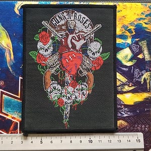 Op de afbeelding: Zwarte patch met het Guns N' Roses logo, een hart, pistolen, rozen en schedels. De patch heeft een zwarte rand en is ongeveer 13 cm breed en 18 cm hoog.