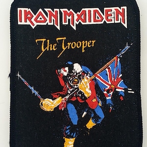 Op de afbeelding: Zwarte strijkpatch met de bandnaam "Iron Maiden" en de titel van het nummer "The Trooper" in witte letters. De patch toont een kleurrijke illustratie van een soldaat die een vlag en een zwaard vasthoudt.