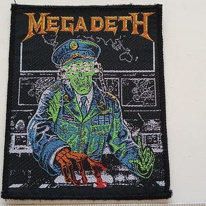 Pode incluir: Um remendo bordado preto e verde com um soldado com rosto de caveira em uniforme militar com o texto "Megadeth" em letras amarelas e laranja.