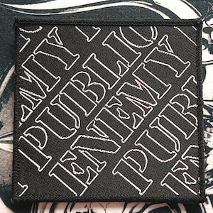 May include: Square black patch with white text that reads "PUBLIC ENEMY" in a repeating pattern. The patch has a black border and is likely an iron-on or sew-on patch for clothing or accessories.