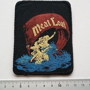 Może przedstawiać: Czarna łatka z tkaniny z czerwono-białym banerem z napisem "Meat Loaf". Baner jest trzymany przez postać w środku łatki.