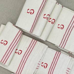 May include: A collection of folded white linen towels with red striped borders and the letter "S" embroidered in red. The towels are neatly stacked, showcasing a classic, vintage aesthetic.