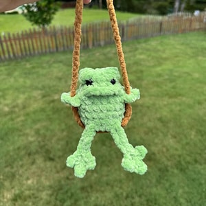 May include: A light green crocheted frog plush toy with black bead eyes, suspended by a brown yarn loop. The frog has a textured, soft yarn surface, a wide mouth, and long dangling legs, crafted from thick, fuzzy yarn in a handmade style.