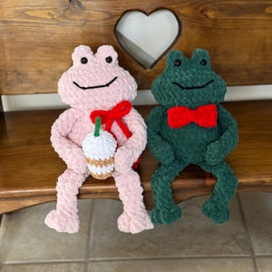 May include: Two plush frog toys. One is pink and holds a white and brown drink with a green straw and a red bow. The other is green with a red bow tie. Both have black eyes and are smiling.