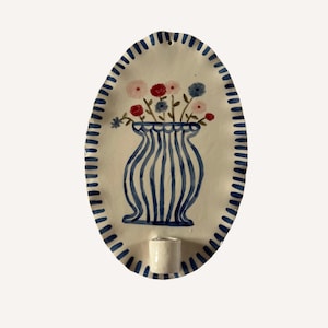 May include: An oval-shaped ceramic wall sconce featuring a hand-painted blue and white striped vase filled with small red, pink, and blue flowers. The edge of the sconce is decorated with blue dashes, and a small cylindrical candle holder is attached.