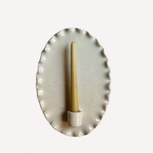May include: A speckled, off-white ceramic candle holder with a scalloped edge. A slender, beige candle is inserted into the holder. The candle holder is oval-shaped and has a minimalist design, suitable for home decor.