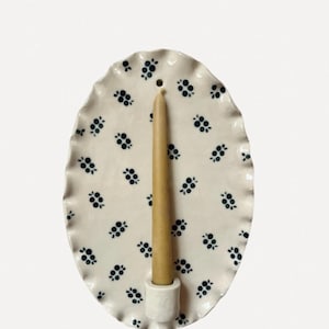 May include: An oval ceramic candle holder in white, with a scalloped edge. It features a repeating pattern of navy blue floral clusters. A beige candle is inserted into the holder.