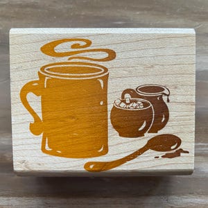 May include: A wooden stamp with a coffee theme. The stamp features a large mug with steam, a small bowl with sugar cubes, a creamer, and a spoon. The design is in a warm brown color.