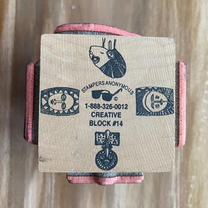 May include: A wooden stamp block with the text "Stampers Anonymous" and "Creative Block #14". The stamp has four decorative designs and a pink rubber base.