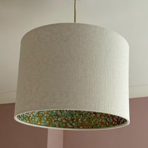 Op de afbeelding: Een cilindrische lampenkap hangt aan het plafond. De buitenkant is gemaakt van lichtbeige stof, terwijl de binnenkant een bloemenpatroon heeft met groene en gele accenten. De lampenkap wordt opgehangen door een beige snoer.
