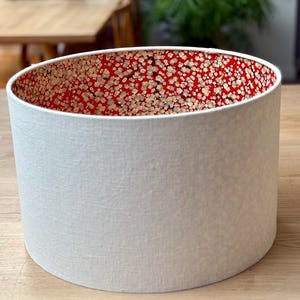 May include: A cylindrical lampshade with a textured white exterior and a red interior patterned with small, gold-coloured floral designs. The lampshade is a home decor item, likely for a table or floor lamp.