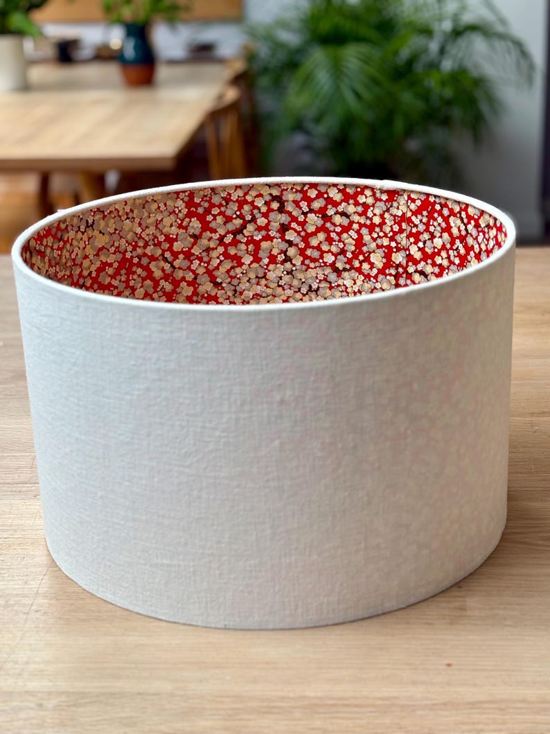 May include: A cylindrical lampshade with a textured white exterior and a red interior patterned with small, gold-coloured floral designs. The lampshade is a home decor item, likely for a table or floor lamp.