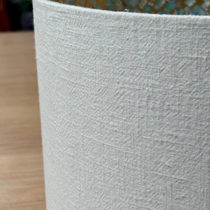 Op de afbeelding: Close-up van een cilindrische lampenkap met een getextureerde, gebroken witte buitenkant. De binnenkant heeft een goud- en turquoise bloemenpatroon. Geschikt voor een tafellamp of vloerlamp.