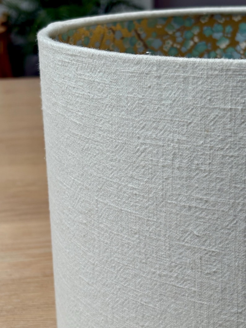 Op de afbeelding: Close-up van een cilindrische lampenkap met een getextureerde, gebroken witte buitenkant. De binnenkant heeft een goud- en turquoise bloemenpatroon. Geschikt voor een tafellamp of vloerlamp.