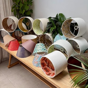 Op de afbeelding: Een verzameling lampenkappen in verschillende maten en patronen. De lampenkappen worden getoond op een houten oppervlak, met een verscheidenheid aan kleuren en texturen, waaronder bloemen-, geometrische en effen ontwerpen. Sommige zijn cilindrisch, andere conisch.