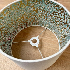 Op de afbeelding: Een cr&egrave;mekleurige lampenkap met een bloemenpatroon aan de binnenkant. De binnenkant heeft een gouden achtergrond met kleine blauwe en witte bloemen. De lampenkap heeft een witte rand en een metalen frame.