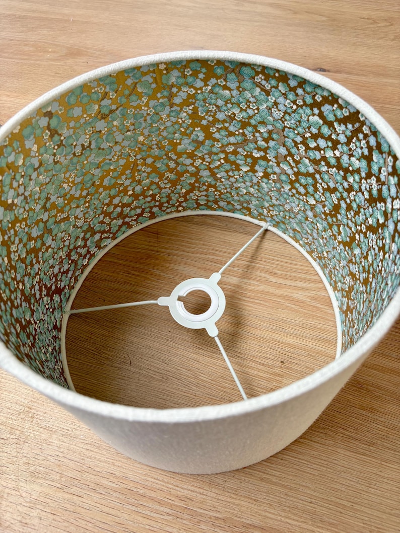 Op de afbeelding: Een cr&egrave;mekleurige lampenkap met een bloemenpatroon aan de binnenkant. De binnenkant heeft een gouden achtergrond met kleine blauwe en witte bloemen. De lampenkap heeft een witte rand en een metalen frame.