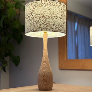 Op de afbeelding: Een houten tafellamp met een patroon lampenkap. De basis is gemaakt van lichtgekleurd hout met een uniek nerfpatroon. De lampenkap heeft een bloemenontwerp in groen- en wittinten. De lamp staat op een houten tafel.