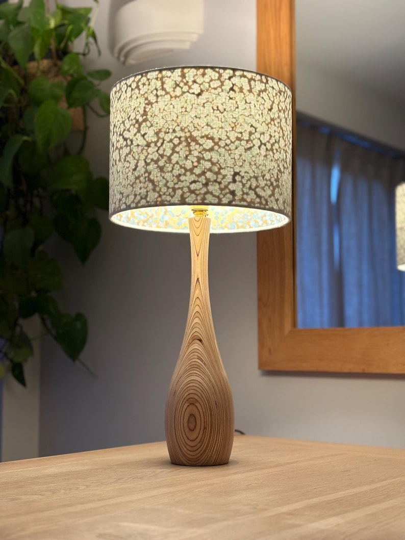 Op de afbeelding: Een houten tafellamp met een patroon lampenkap. De basis is gemaakt van lichtgekleurd hout met een uniek nerfpatroon. De lampenkap heeft een bloemenontwerp in groen- en wittinten. De lamp staat op een houten tafel.