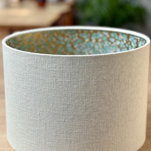 Op de afbeelding: Cilindrische lampenkap met een getextureerde, gebroken witte buitenkant. De binnenkant heeft een turquoise en goud gespikkeld patroon. De lampenkap is ontworpen om licht te verspreiden en een decoratief element aan een lamp toe te voegen.