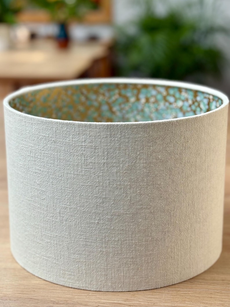 Op de afbeelding: Cilindrische lampenkap met een getextureerde, gebroken witte buitenkant. De binnenkant heeft een turquoise en goud gespikkeld patroon. De lampenkap is ontworpen om licht te verspreiden en een decoratief element aan een lamp toe te voegen.