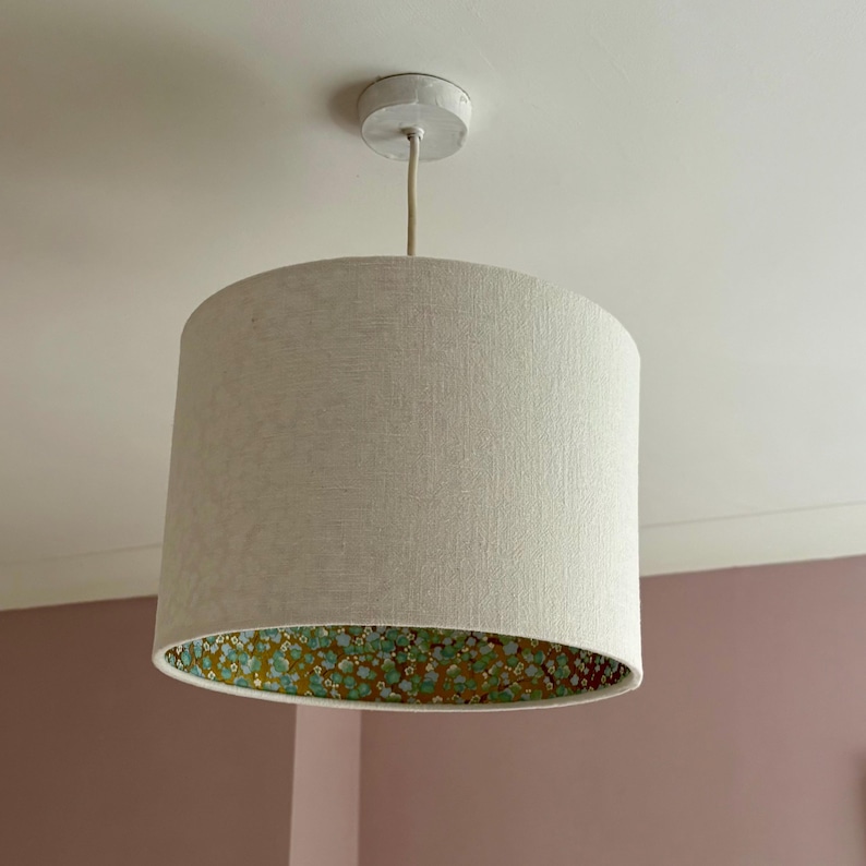 Op de afbeelding: Een cilindrische lampenkap hangt aan het plafond. De buitenkant is gemaakt van lichtbeige stof, terwijl de binnenkant een bloemenpatroon heeft met groene en gele accenten. De lampenkap wordt opgehangen door een beige snoer.
