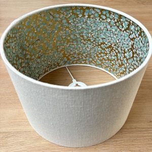 Op de afbeelding: Cilindrische lampenkap met een cr&egrave;mekleurige buitenkant en een bloemmotief aan de binnenkant. De binnenkant heeft een gouden achtergrond met kleine blauwe en witte bloemclusters. De lampenkap is ontworpen voor een armatuur.