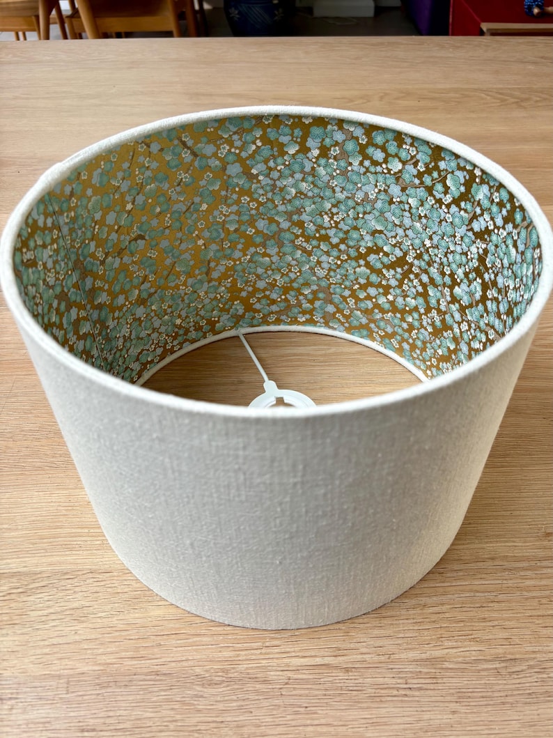 Op de afbeelding: Cilindrische lampenkap met een cr&egrave;mekleurige buitenkant en een bloemmotief aan de binnenkant. De binnenkant heeft een gouden achtergrond met kleine blauwe en witte bloemclusters. De lampenkap is ontworpen voor een armatuur.