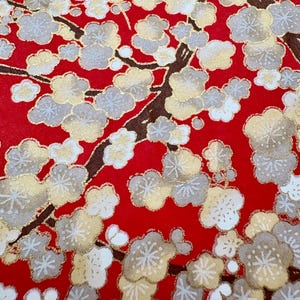 May include: A close-up of red fabric with a floral pattern. The design features white, gold, and grey blossoms on brown branches. A white cord is visible at the top, suggesting a bag or textile item.