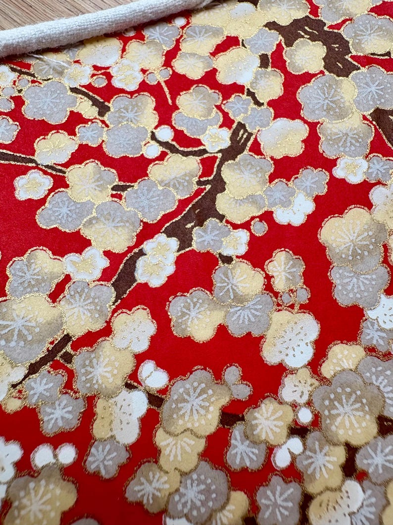 May include: A close-up of red fabric with a floral pattern. The design features white, gold, and grey blossoms on brown branches. A white cord is visible at the top, suggesting a bag or textile item.