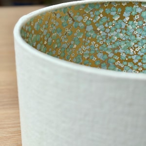 Op de afbeelding: Witte cilindrische lampenkap met een getextureerd oppervlak. De binnenkant heeft een bloemenpatroon met blauwe en witte bloemen op een gouden achtergrond. De lampenkap is ontworpen om licht te verspreiden.