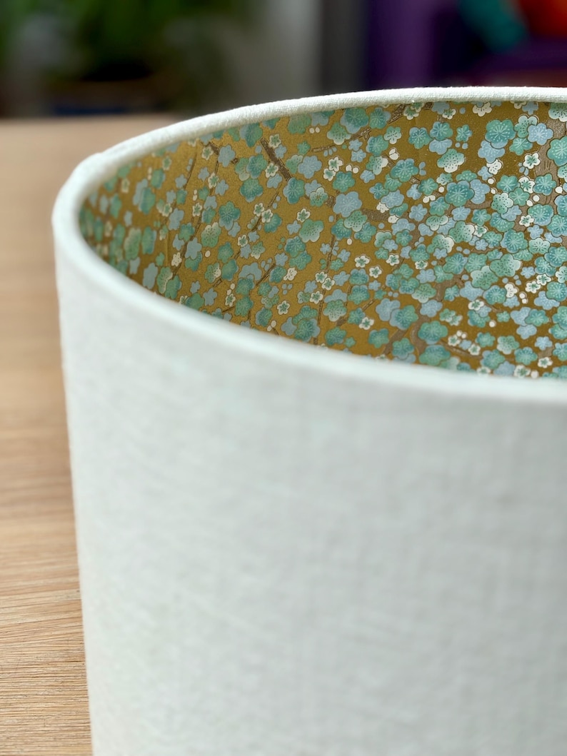 Op de afbeelding: Witte cilindrische lampenkap met een getextureerd oppervlak. De binnenkant heeft een bloemenpatroon met blauwe en witte bloemen op een gouden achtergrond. De lampenkap is ontworpen om licht te verspreiden.