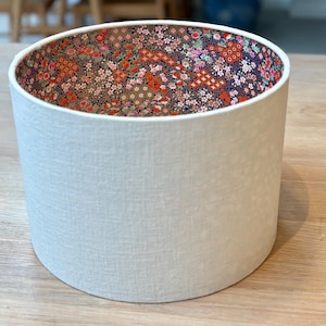 Puede incluir: Una pantalla de lámpara cilíndrica con un exterior texturizado de color blanco roto. El interior presenta un colorido estampado floral con flores rojas, rosas y verdes sobre un fondo oscuro. La pantalla está diseñada para difundir la luz.